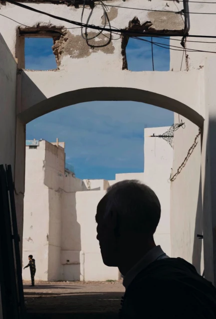 Silhouette of a grey-haired adult in a white stone archway in an outdoor setting, with another human figure in the distance.