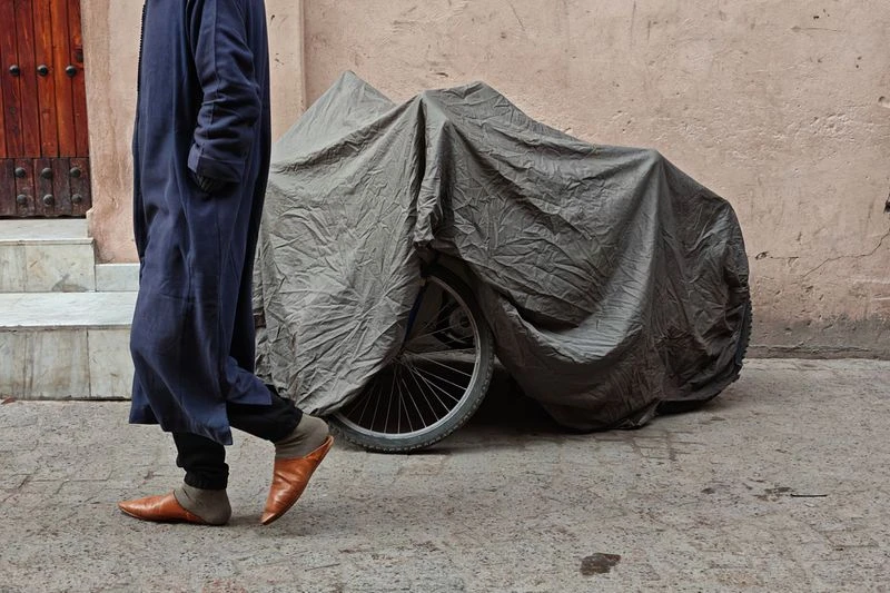 Person in traditional blue robe walking past a bicycle covered with a grey tarp, set against a light-orange building background.