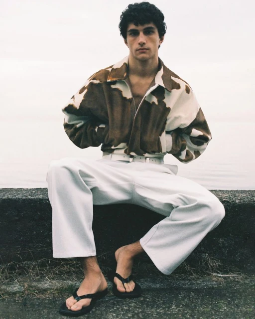 A young adult male sits on a concrete wall in an outdoor setting, wearing an animal print jacket, wide leg white pants and flip flops.