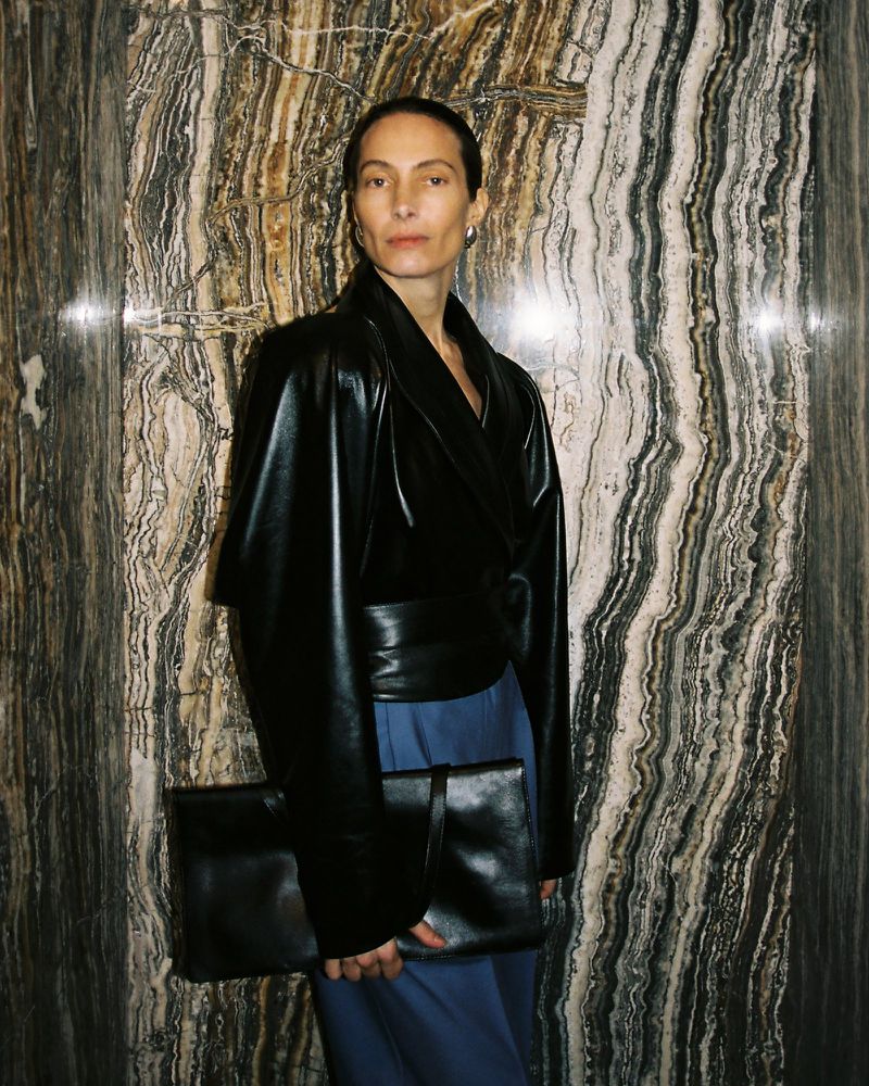 Adult woman wearing a black jacket and blue skirt poses indoors with a large black bag in front of a marble wall.