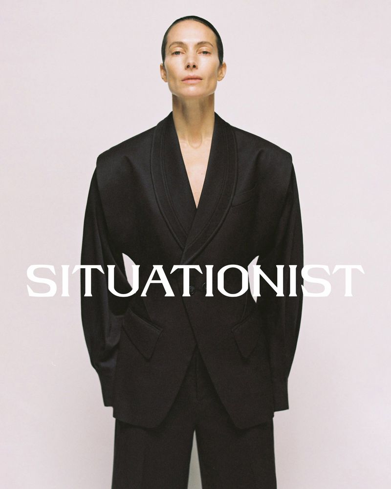 Woman poses indoors wearing a black oversized blazer and black pants against a neutral background with 'SITUATIONIST' text overlay.