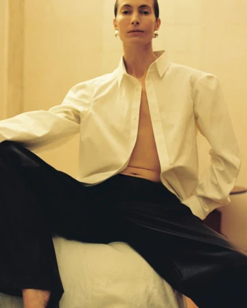 A woman wearing a white button-down shirt and black pants is sitting indoors with a neutral background, posing for a fashion shot.