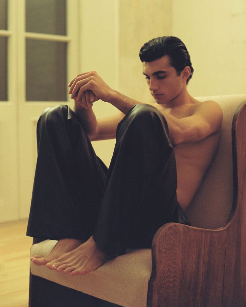 Bare-chested man sitting in a chair with his hands clasped and gazing downward in an indoor setting with a window in the background.