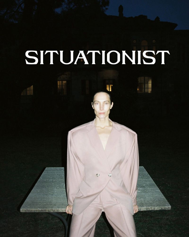 A person in a suit sits on a concrete table in front of a house at night, with the word 'SITUATIONIST' in bold, white letters above them.