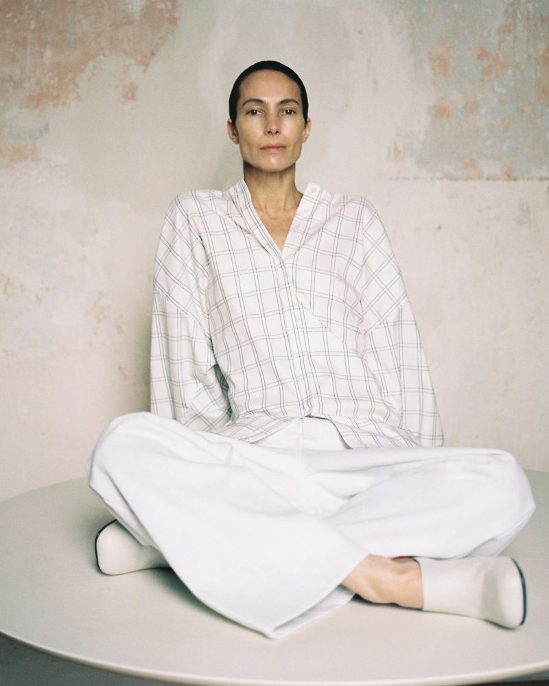 Adult woman sits cross-legged on a white table wearing a white plaid shirt and white pants against a textured neutral wall in a studio.