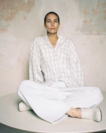 Adult woman sits cross-legged on a white table wearing a white plaid shirt and white pants against a textured neutral wall in a studio.