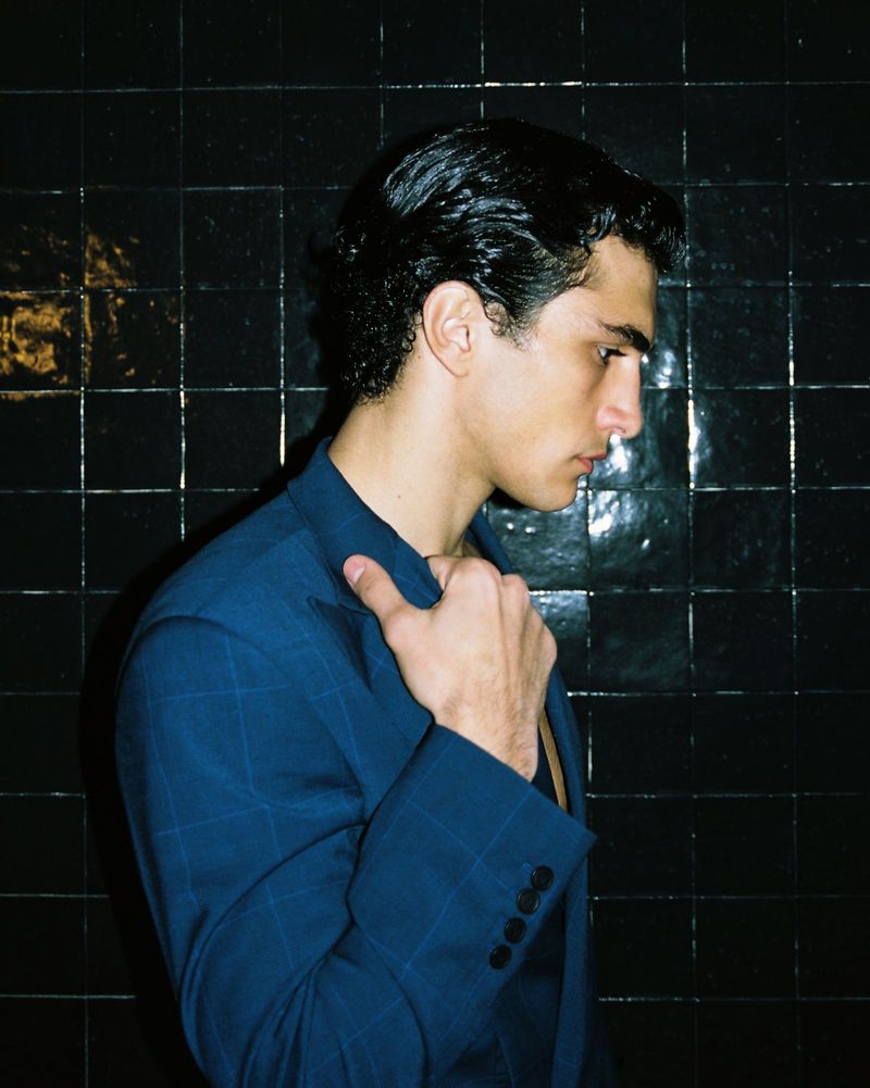 Portrait of a young adult male in a blue patterned suit jacket against a dark tiled background, looking off to the side in a studio.
