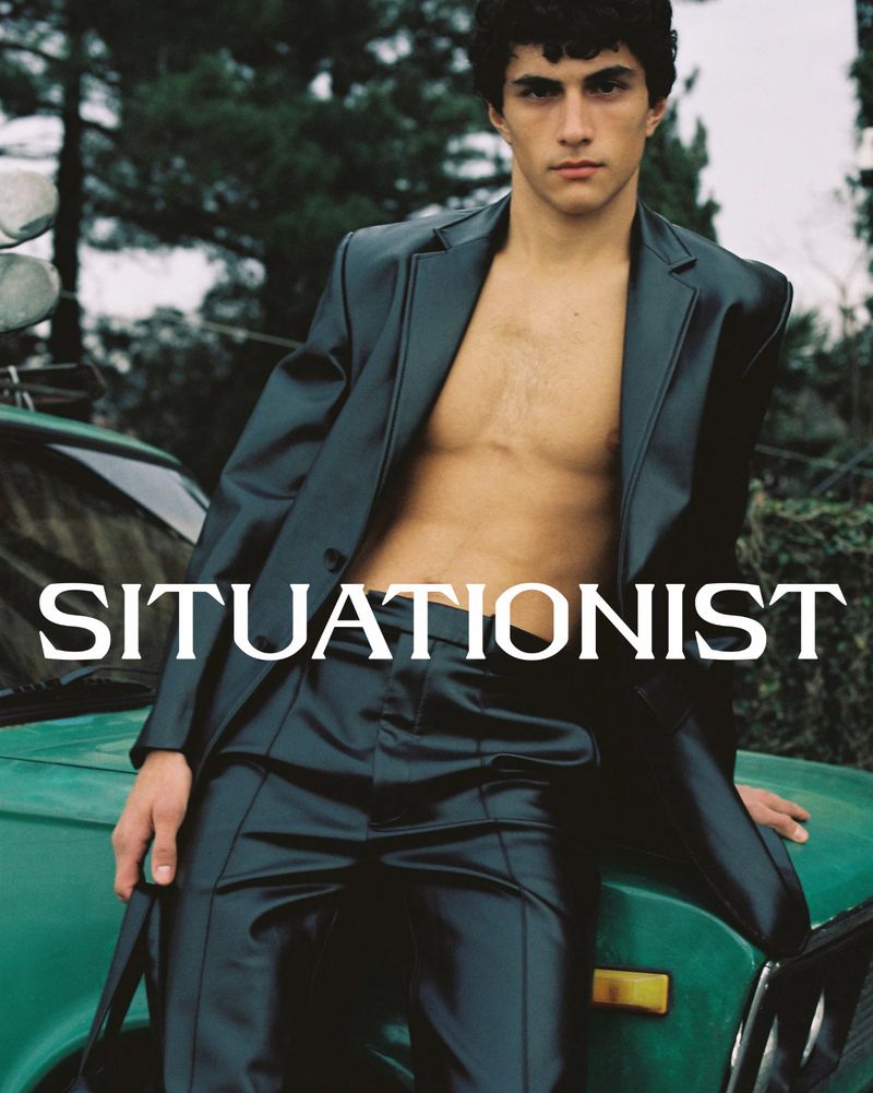 Fashion portrait of a shirtless man wearing a suit, leaning against a green car in an outdoor setting with natural lighting.