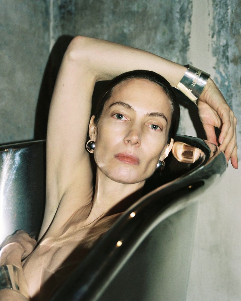 Adult woman with silver cuff bracelets and ball earrings poses inside a silver-colored bathtub against a textured concrete wall.