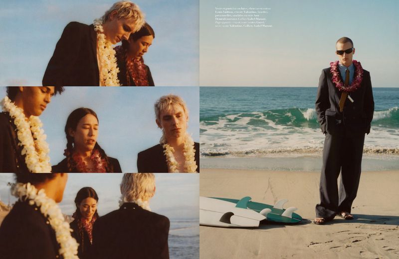 Three young adults in suits wearing floral leis on a beach, with a single male standing near surfboards in the background.
