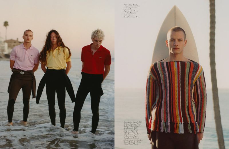 Editorial shot of three people in polo shirts and wetsuit pants on the beach, and a person with a surfboard by the ocean.