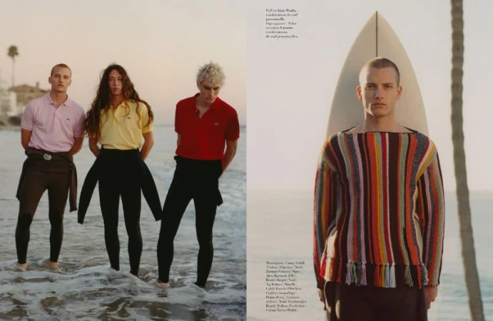 Editorial shot of three people in polo shirts and wetsuit pants on the beach, and a person with a surfboard by the ocean.