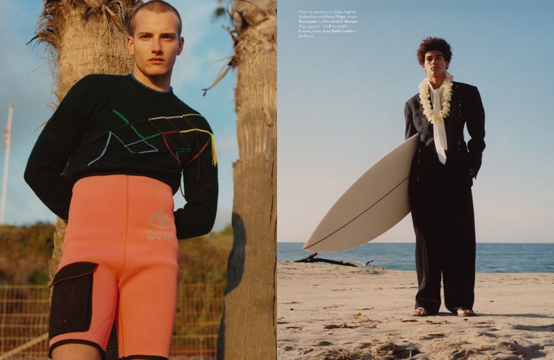 Two male models pose on a beach, one with a surfboard, against a blue sky. Beach fashion photo shoot.