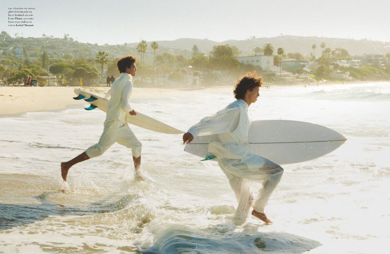 Two young barefoot men in light-colored linen clothing run in ocean surf carrying surfboards on a sunny day with beach and buildings.