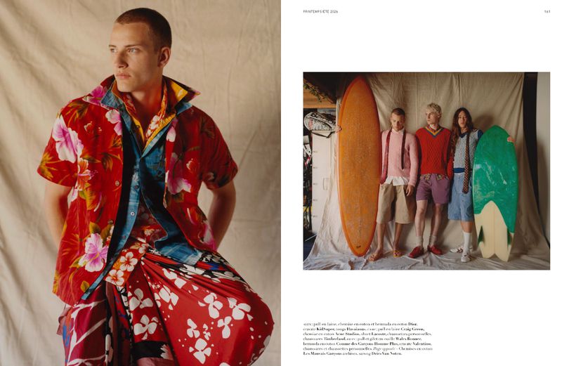 Editorial fashion image: A man in a colorful floral shirt and skirt stands next to a group with surfboards in front of a cream backdrop.