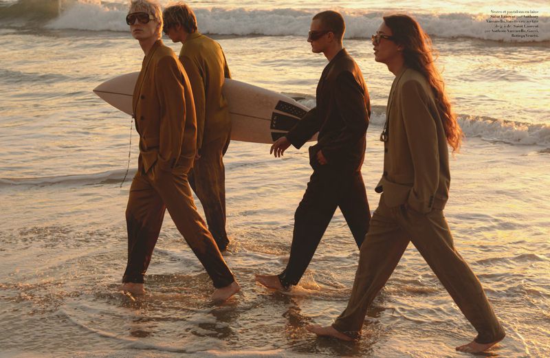 Four young adult models walk barefoot through ocean water at sunset, wearing fashionable brown and olive suits while one carries a surfboard.