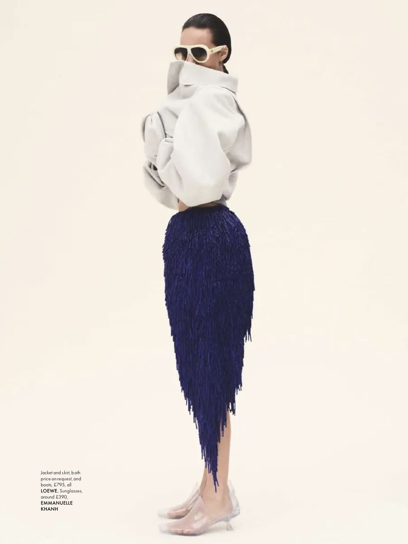 Fashion editorial featuring a model in a voluminous white top and blue fringe skirt, clear shoes against an off-white background.