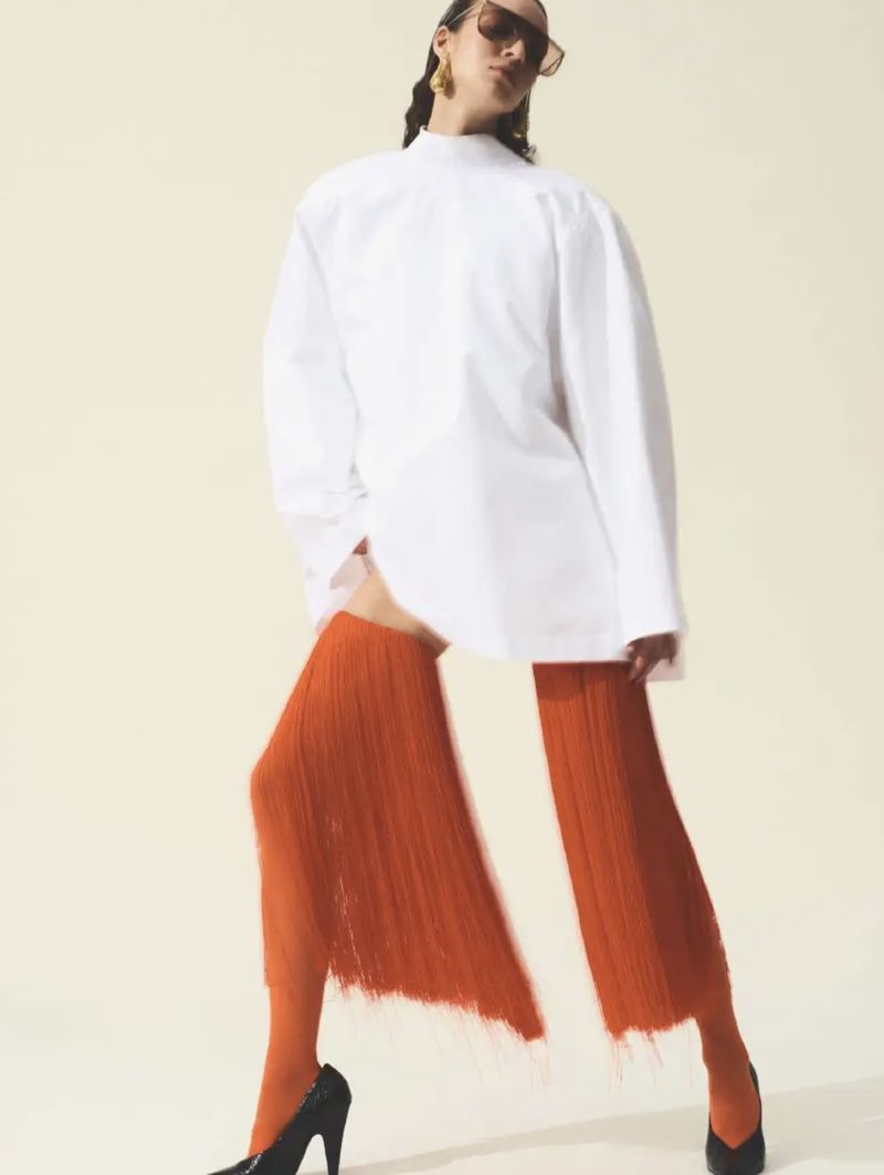A slender fashion model poses in a white oversized shirt, orange textured leg coverings, and black heels against a neutral backdrop.