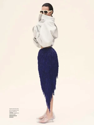 Model Emm Arruda wearing an oversized white jacket and blue fringed skirt against a white studio background.