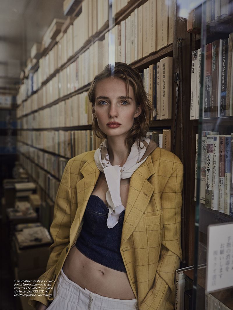 Woman in yellow blazer and blue top poses in front of bookshelf, showcasing fashion and editorial style with natural light indoors.