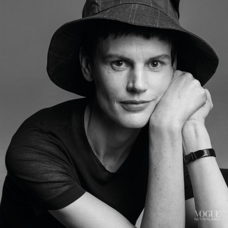 Monochrome portrait of a person with short hair wearing a hat, with hands clasped beneath their chin, in front of a solid grey backdrop.