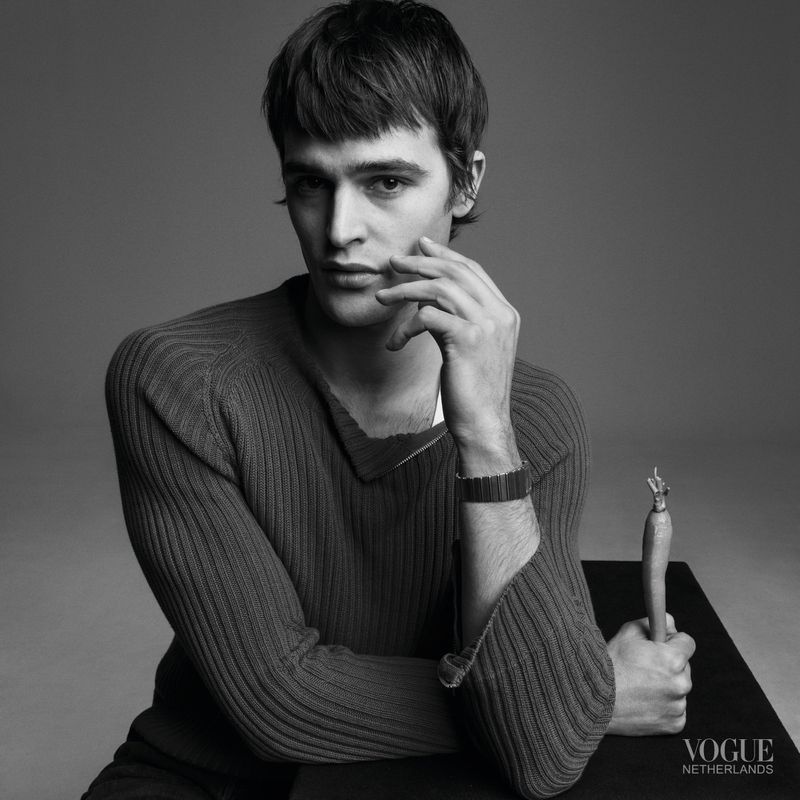 Black and white portrait of a young adult male in a sweater, hand to face, with a sculpture on a black surface. Fashion editorial.