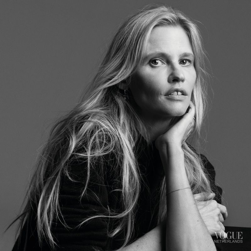 Black and white portrait of a fair-skinned woman with long blonde hair, wearing a black turtleneck, posed in front of a gray backdrop.