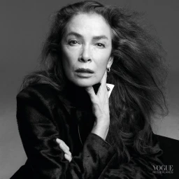 Portrait of an older woman with long dark hair wearing a black top against a grey seamless background for Vogue Netherlands