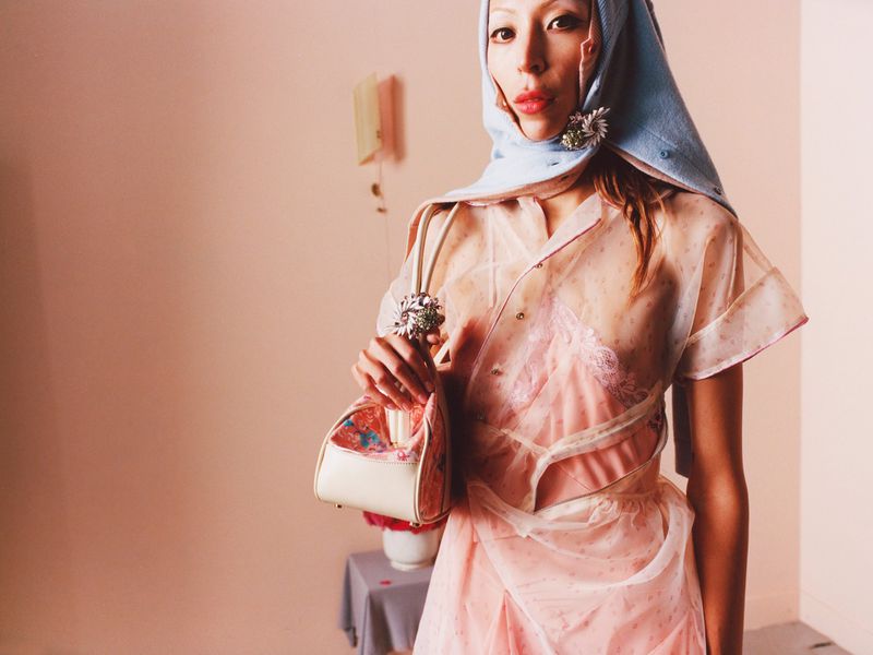 Woman in sheer shirt, wrapped headscarf, and handbag poses in front of a peach wall with a vase and flower on a table.