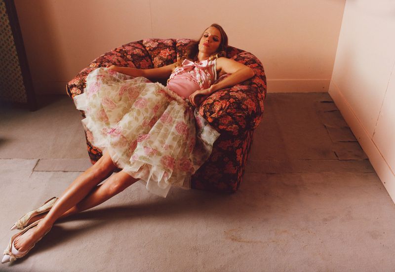 Woman in pink top and floral skirt poses in a floral armchair, wearing matching shoes, in a photo studio with pink walls.