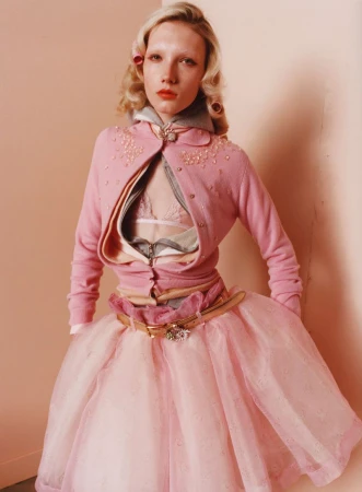 Blonde woman in pink cardigan and layered pink tulle skirt poses against a peach wall, fashion portrait, studio lighting, high angle.