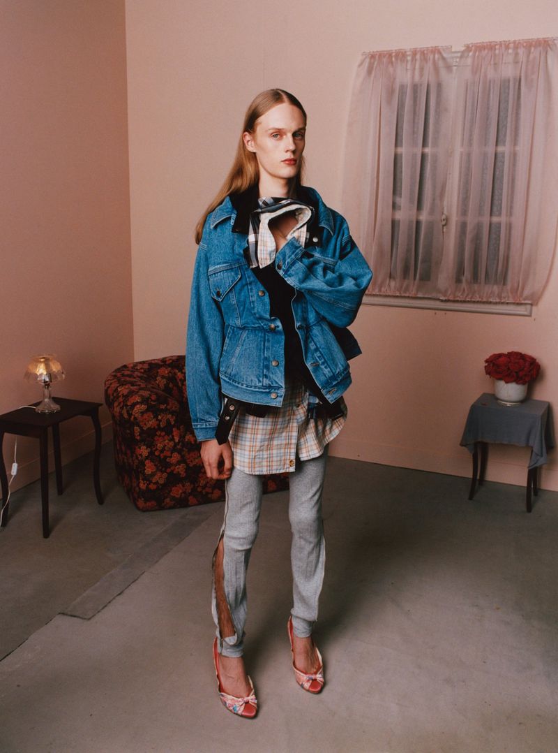 Fashion shot of a person in denim jacket, layered tops, distressed jeans, and floral heels in an indoor setting with pale pink walls.