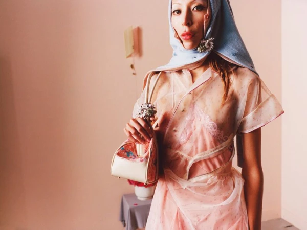 Model in pink dress and blue headscarf holding a handbag against a plain pink wall for August Barron Real Housewife project