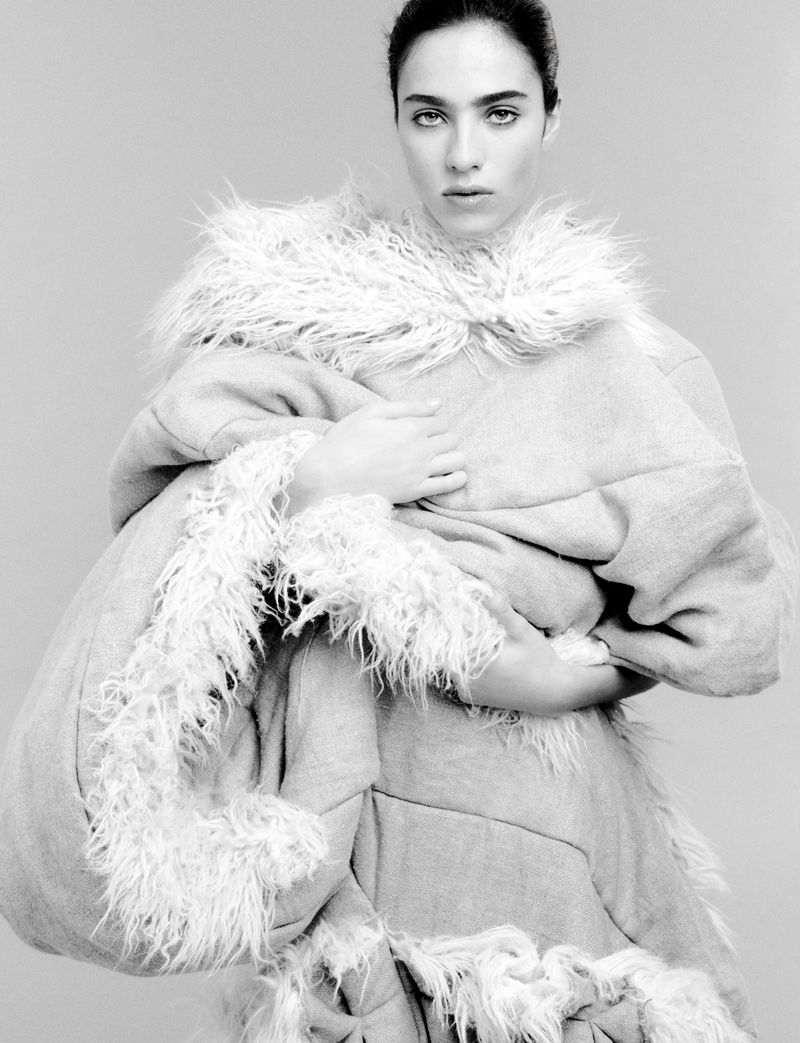 Monochrome portrait of a woman in a sculptural coat with a furry collar, embracing herself in a studio setting against a gray backdrop.