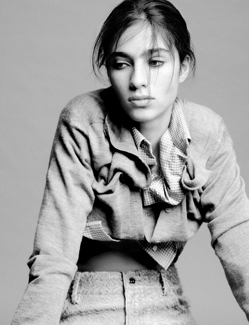 Monochrome portrait of a young adult female with a neutral expression, wearing a cropped jacket and patterned shirt against a grey backdrop.
