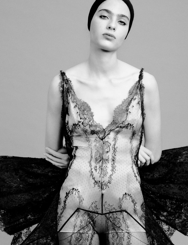 Monochrome fashion portrait of a woman wearing a lace slip dress and a cap, set against a grey studio backdrop in soft lighting.