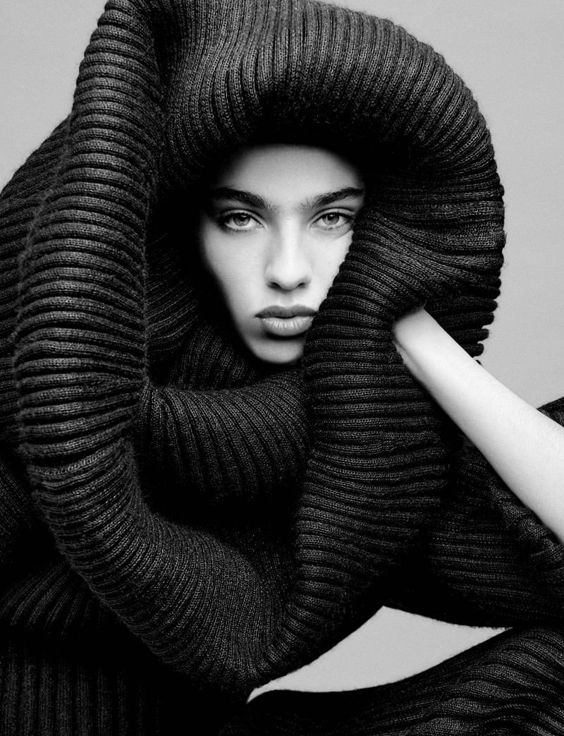 Monochrome portrait of a woman with dark hair and dramatic makeup, wrapped in a thick, ribbed knit fabric, posing in a photo studio.