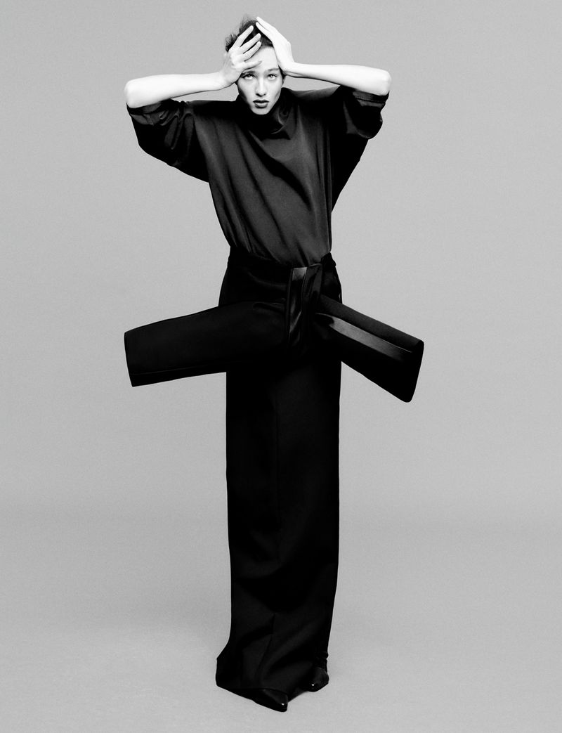 Fashion photo of a slender woman in a black high-fashion outfit, standing in a studio against a grey backdrop, hands raised to her head.