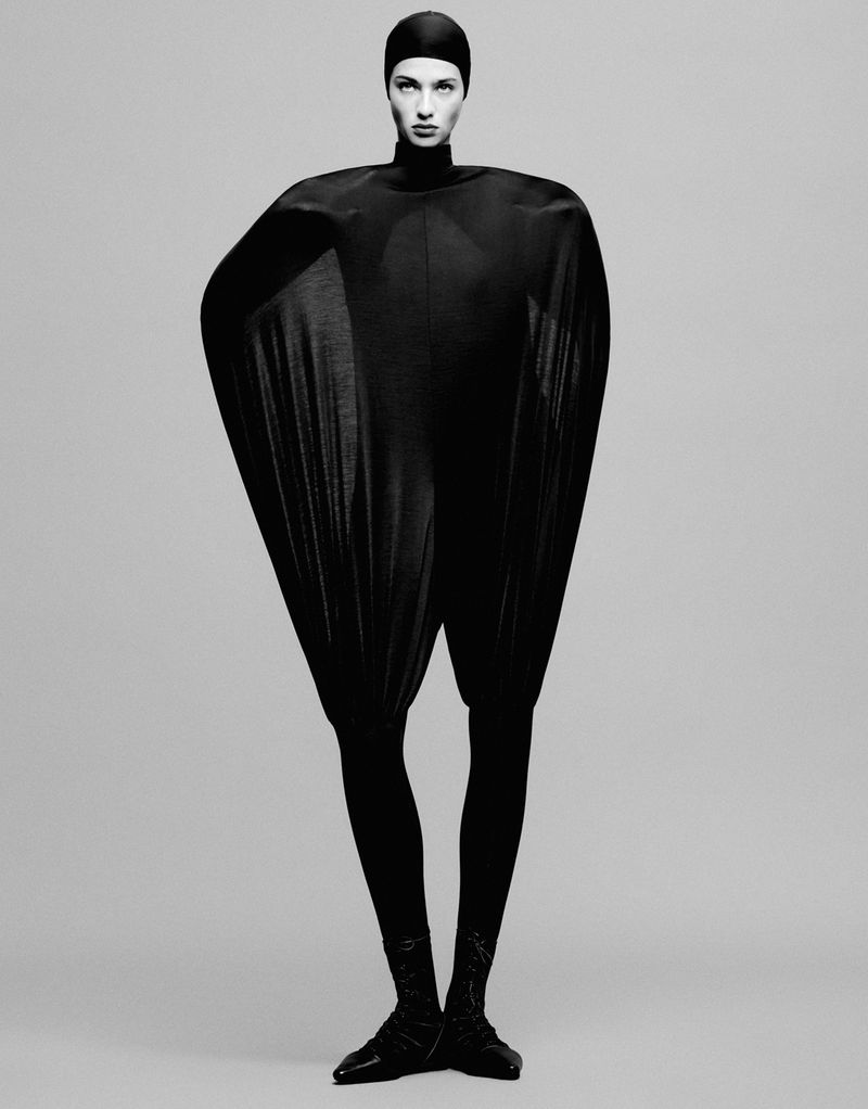 Full-length fashion shot of adult female model in black futuristic catsuit against grey backdrop, eyes forward, arms at side, studio lighting.