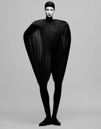 Full-length fashion shot of adult female model in black futuristic catsuit against grey backdrop, eyes forward, arms at side, studio lighting.