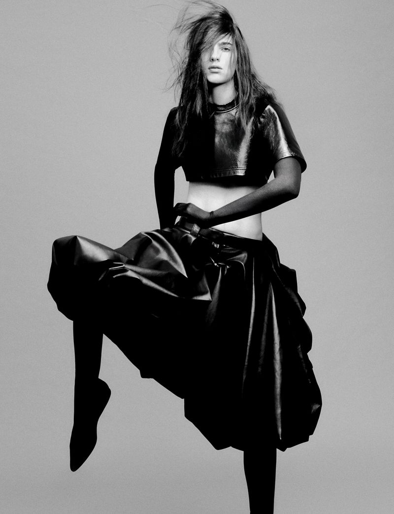Fashion editorial shot of a young adult female model in a black, shiny crop top and voluminous skirt, with wind-blown hair, against a gray backdrop.