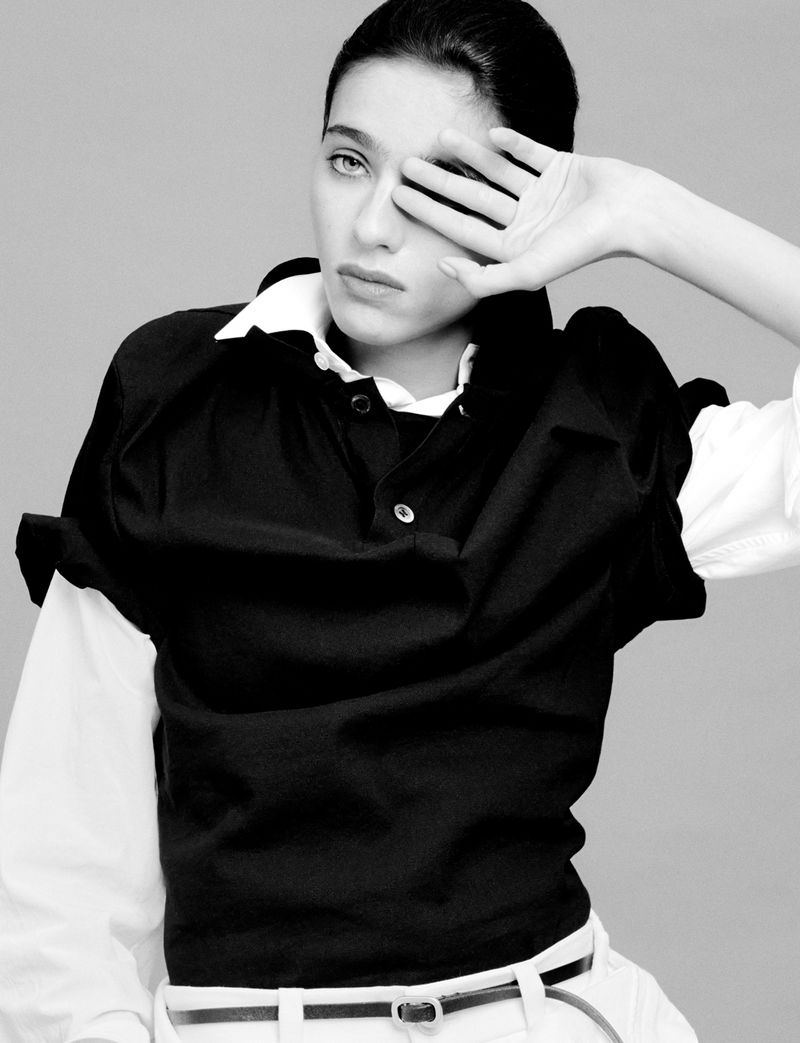 Monochrome portrait of an adult woman covering one eye with her hand, wearing a dark top and light-colored pants against a grey backdrop.