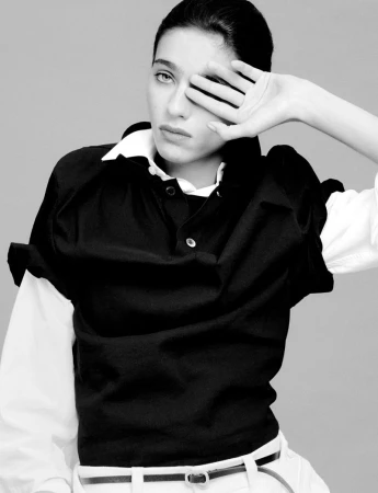 Monochrome portrait of an adult woman covering one eye with her hand, wearing a dark top and light-colored pants against a grey backdrop.