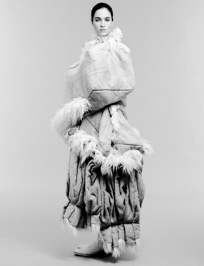 Monochrome fashion portrait of a model wearing an oversized, padded, avant-garde coat with fur trim in a minimalist photo studio.