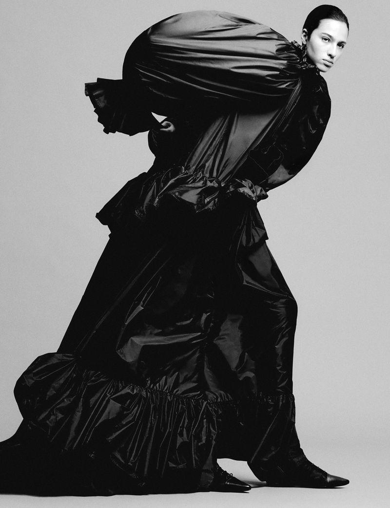 Monochrome fashion shot: an adult woman in a dramatic black gown poses against a neutral backdrop, showcasing a unique ruffled design.