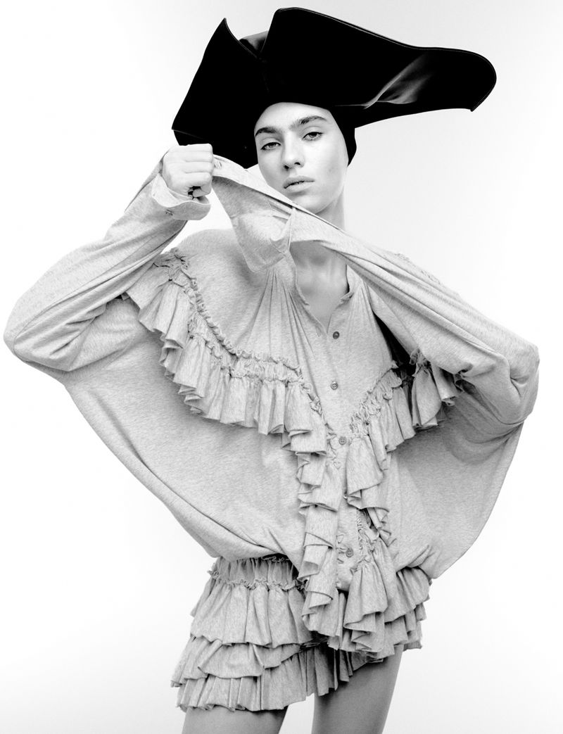 Monochrome studio shot of a person in gray ruffled outfit and oversized black hat. The person is holding the outfit's fabric.