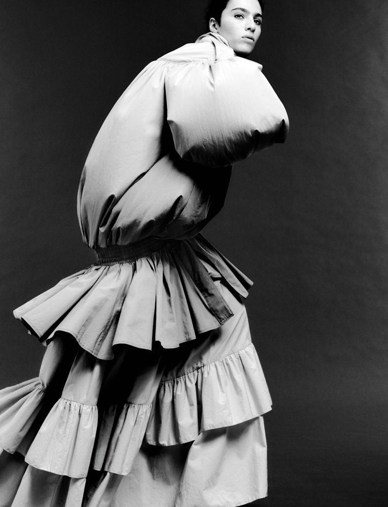 Fashion shot of an adult female model posing in a large, layered ruffle dress with a voluminous puffy top against a grey studio backdrop.