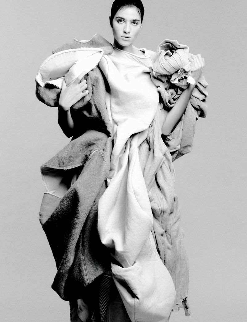 Black and white fashion portrait of a woman wearing clothes of stacked and draped fabric, set against a neutral grey studio backdrop.
