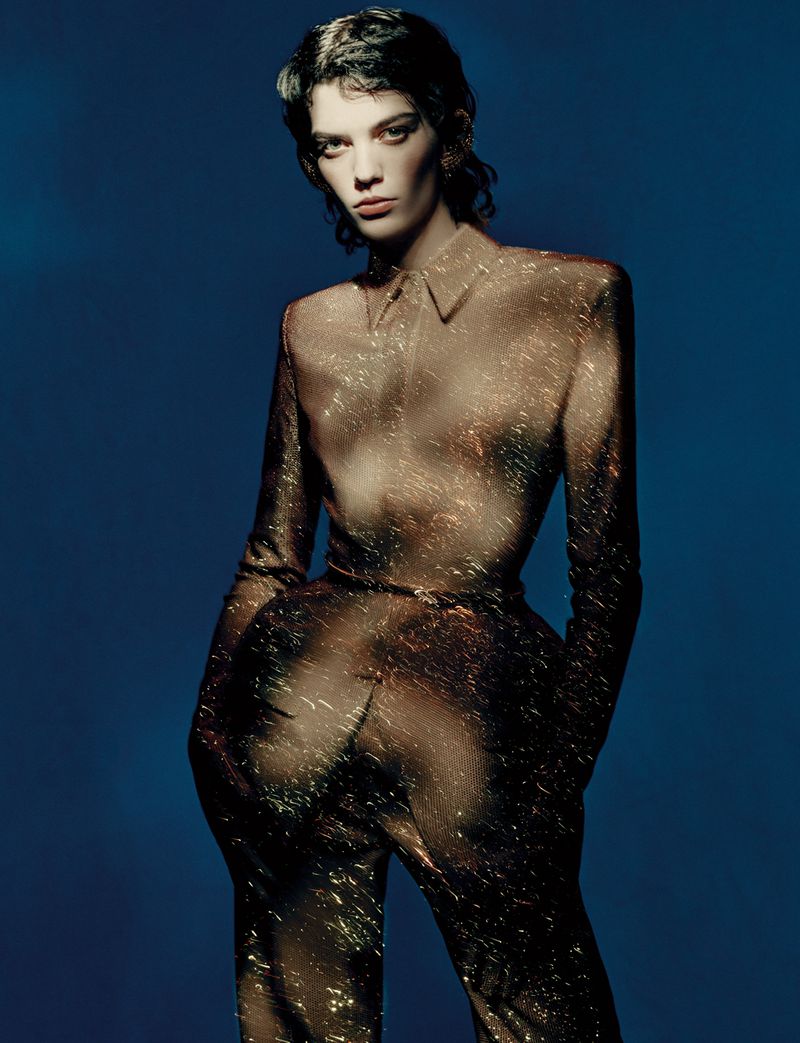 Woman posing in sheer long-sleeved shirt and pants against a blue background, showcasing a fashion-forward look with visible skin.