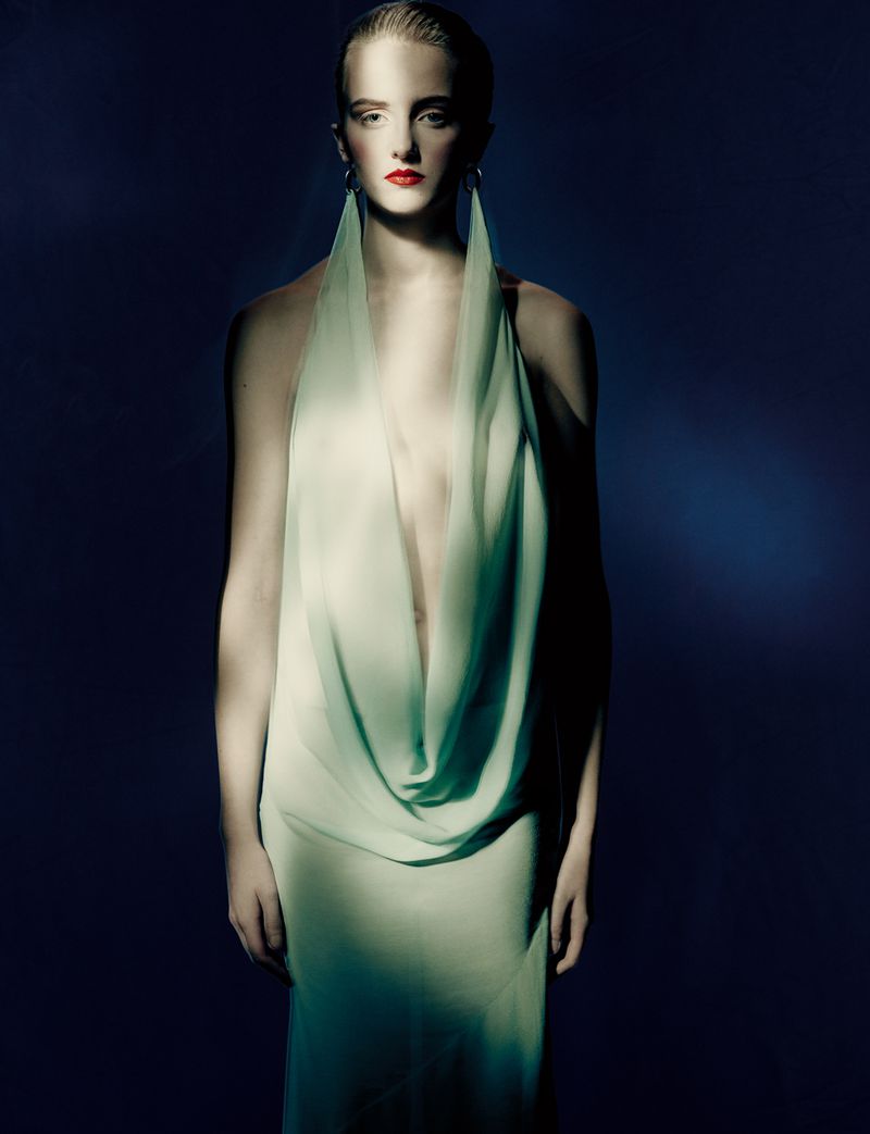 Elegant woman in a light green gown with long earrings stands against a dark blue backdrop in a fashion or beauty shot.